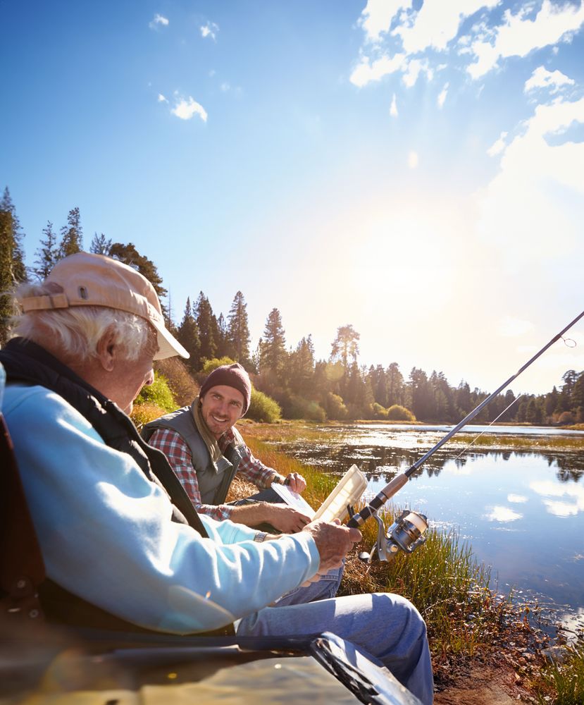 Bild på en äldre man som fiskar med en yngre man vid en sjö i skogen. Solen skiner.