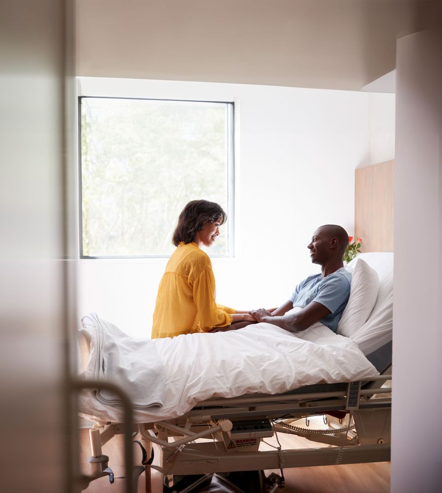 En person besöker sin partner som ligger i en sjukhussäng.