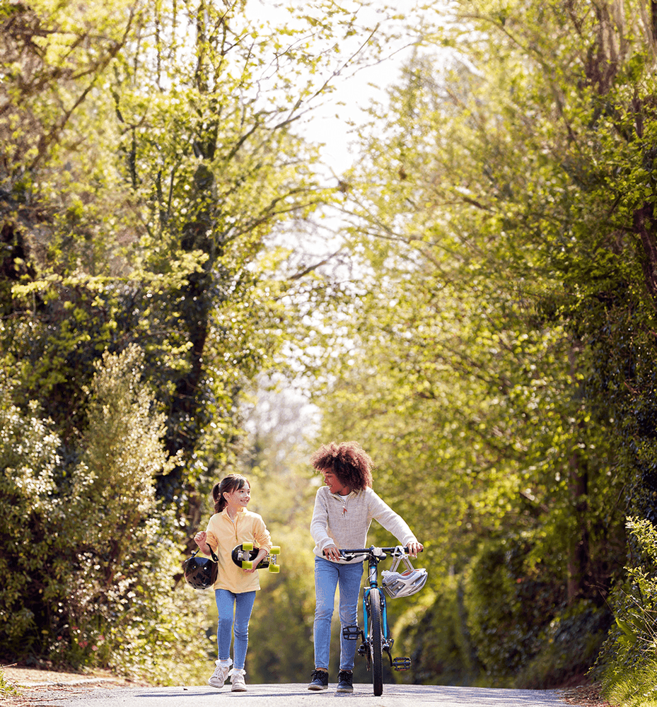 Två barn går och pratar. En leder en cykel och en bär på en skateboard.